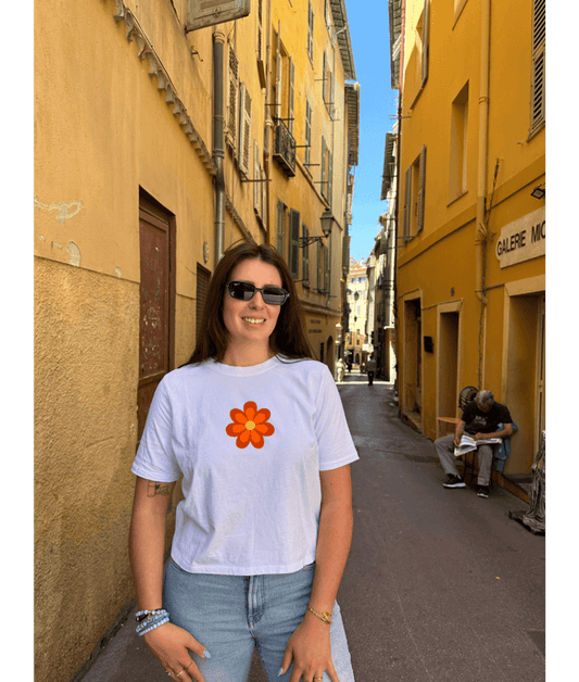 Crop top blanc court, manche courte, 100% coton. Personnalisation du crop top grâce à une technique de flocage, le DTF, pour une meilleure qualité d’impression et de tenue. La personnalisation est floquée à l’avant au niveau du cœur, en haut à gauche et à l’arrière en grand dans le dos. Ici, il s’agit de la personnalisation de DTF fleur orange seventies 70. Rue vieux Nice 06