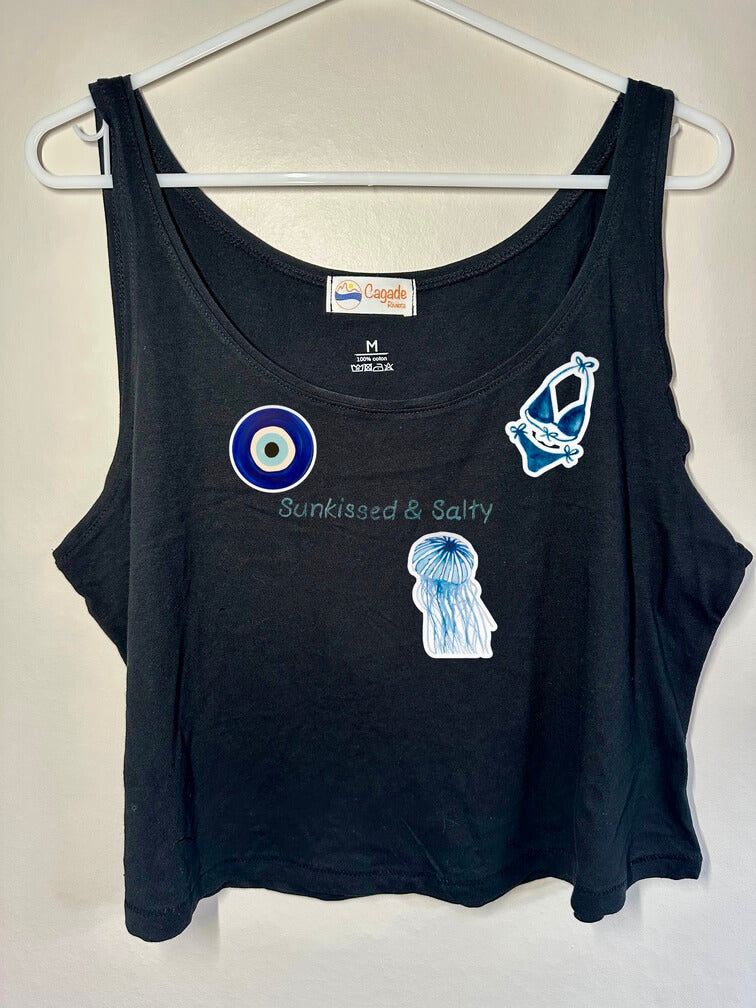 Débardeur noir court, crop top, 100% coton. Personnalisation du débardeur grâce à des broderies et du flocage DTF. Toutes les broderies sont faites à la commande. Ici, il s’agit de la broderie écriture "sunkissed & salty". et flocage DTF bleu de l'oeil de sainte lucie d'un maillot de bain femme bleu et d'une méduse bleue.
