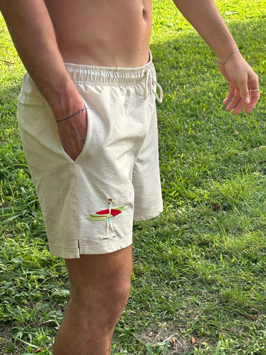 Mannequin homme portant maillot plage short beige zoom sur la broderie surf, surfeur avec sa planche de surf. Décor paradisiaque, Guadeloupe.