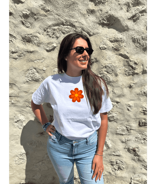 T-shirt oversize blanc, manche courte, 100% coton. Personnalisation du t-shirt grâce à une technique de flocage, le DTF, pour une meilleure qualité d’impression et de tenue. La personnalisation est floquée à l’avant au milieu en haut et à l’arrière en grand dans le dos. Ici, il s’agit de la personnalisation de DTF fleur orange style seventies. Porté par un modèle au bord de l'eau coucher de soleil mer plage été.