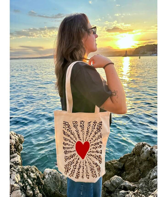 Tote bag, sac de course en coton. Personnalisation du débardeur grâce à une technique de flocage, le DTF, pour une meilleure qualité d’impression et de tenue. La personnalisation est floquée au centre, en haut milieu. Tout est fait à la commande selon la personnalisation choisie. Ici, il s’agit de la personnalisation de DTF, flocage coeur rouge et léopard. Femme sac porté épaule, mer, coucher de soleil.