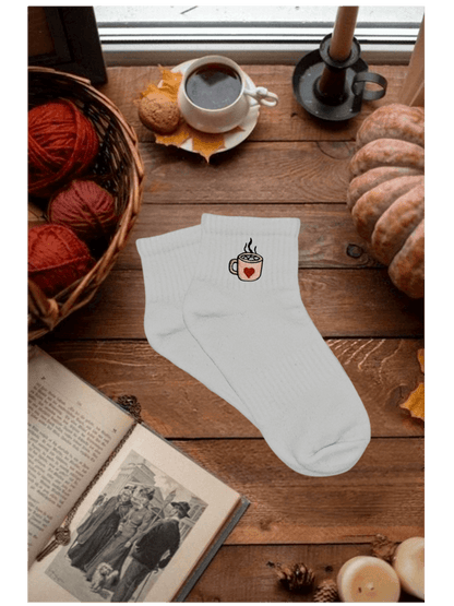 Paire de chaussettes basses blanches en coton. Broderie sur le côté tasse de chocolat chaud rose avec un coeur rouge et des marshmallows. Dans un décor automnal, cozy, citrouille, bougie, livre, pelotte de laine, café. Idée personnalisation broderie automne.