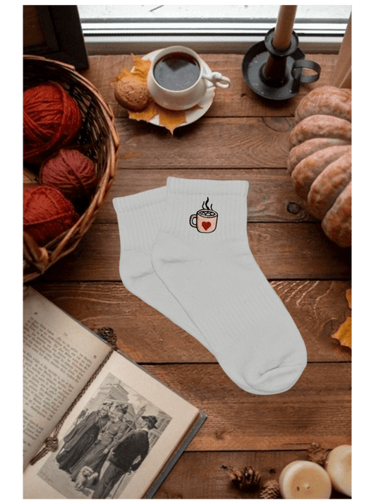 Paire de chaussettes basses blanches en coton. Broderie sur le côté tasse de chocolat chaud rose avec un coeur rouge et des marshmallows. Dans un décor automnal, cozy, citrouille, bougie, livre, pelotte de laine, café. Idée personnalisation broderie automne.