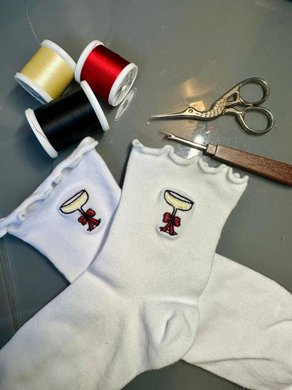 Chaussettes blanches courtes avec petits froufrous à l’extrémité, 100% coton. Personnalisation des chaussettes grâce à des broderies. Il faut choisir parmi un choix de broderies dessinées à la main sur le thème de l’hiver, Noël, fêtes fin d’année, winter. La broderie est présente sur le côté extérieur des chaussettes. Toutes les broderies sont faites à la commande selon la personnalisation choisie. Ici, il s’agit de la broderie coupe de champagne avec noeud rouge