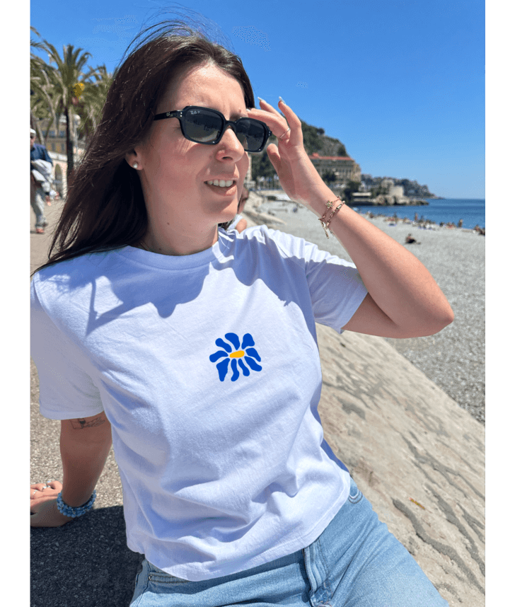 Modèle femme portant un crop top blanc en coton personnalisation flocage DTF fleur bleu à l'avant au centre. Assise sur la promenade des anglais Nice. Plage galets palmier mer