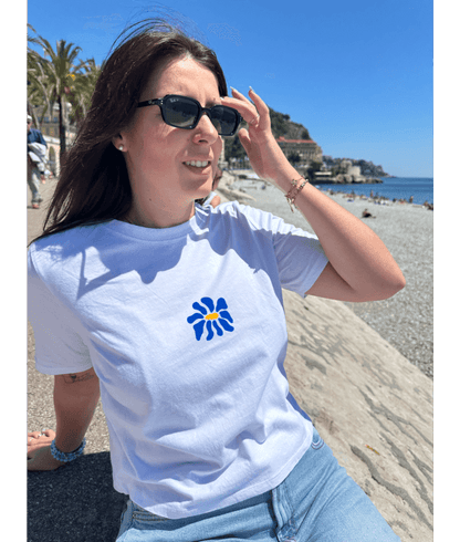 Modèle femme portant un crop top blanc en coton personnalisation flocage DTF fleur bleu à l'avant au centre. Assise sur la promenade des anglais Nice. Plage galets palmier mer