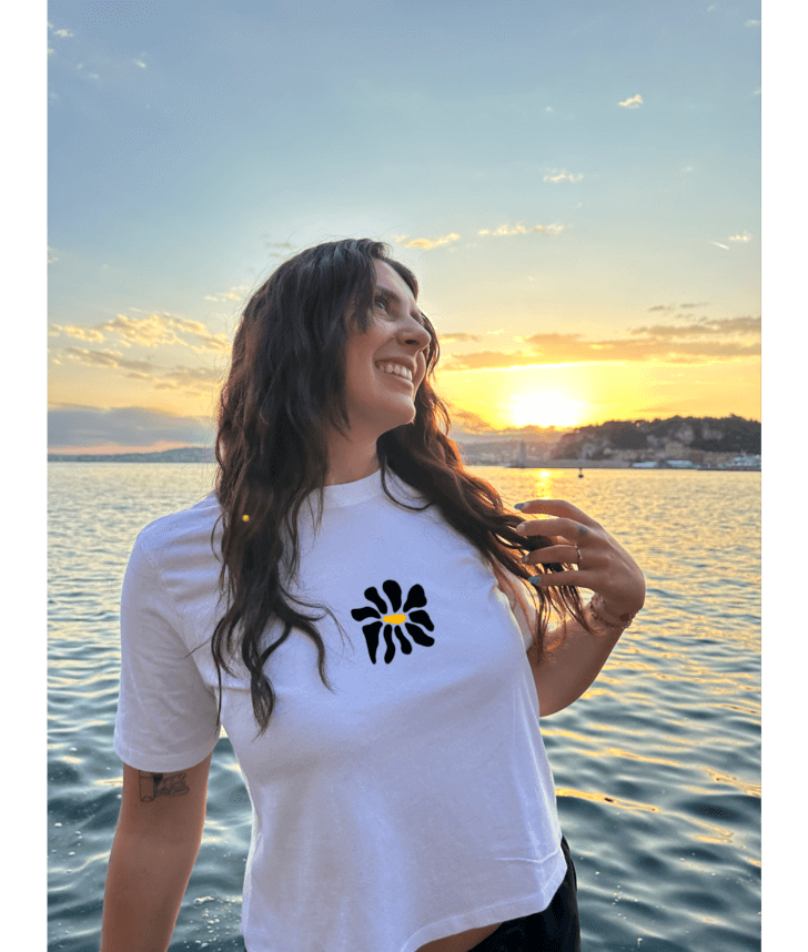 Modèle femme portant un crop top blanc collection summer personnalisation DTF fleur noir devant au centre. Décor plage, mer, coucher de soleil