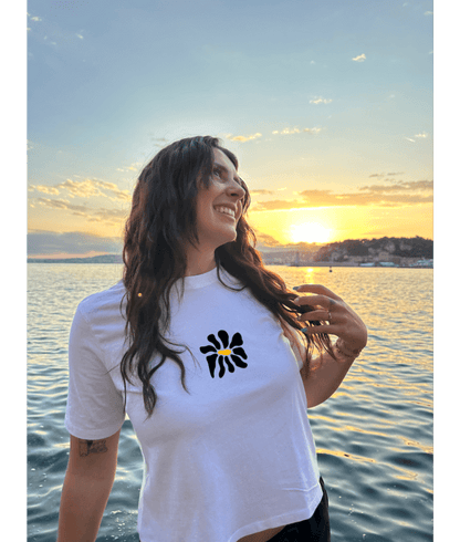 Modèle femme portant un crop top blanc collection summer personnalisation DTF fleur noir devant au centre. Décor plage, mer, coucher de soleil