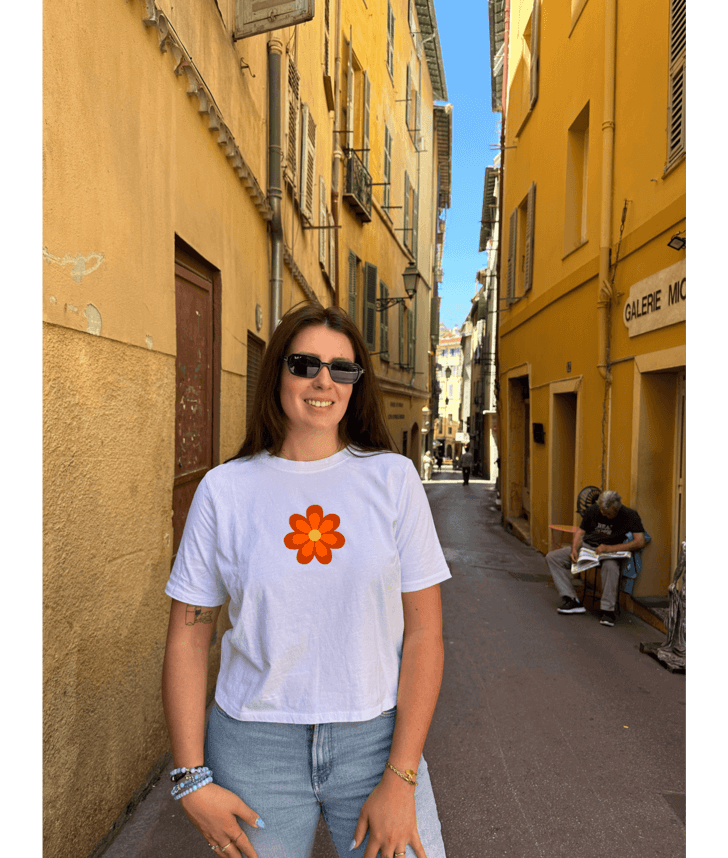 Crop top blanc court, manche courte, 100% coton. Personnalisation du crop top grâce à une technique de flocage, le DTF, pour une meilleure qualité d’impression et de tenue. La personnalisation est floquée à l’avant au niveau du cœur, en haut à gauche et à l’arrière en grand dans le dos. Ici, il s’agit de la personnalisation de DTF fleur orange seventies 70. Rue vieux Nice 06