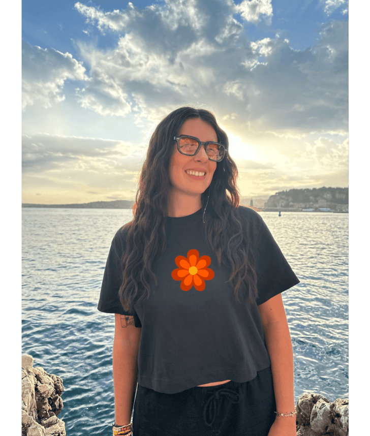 Crop top noir court, manche courte, 100% coton. Personnalisation du crop top grâce à une technique de flocage, le DTF, pour une meilleure qualité d’impression et de tenue. La personnalisation est floquée à l’avant au niveau du cœur, en haut à gauche et à l’arrière en grand dans le dos. Ici, il s’agit de la personnalisation de DTF fleur orange avant seventies 70. 