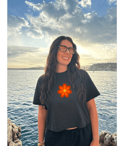 Crop top noir court, manche courte, 100% coton. Personnalisation du crop top grâce à une technique de flocage, le DTF, pour une meilleure qualité d’impression et de tenue. La personnalisation est floquée à l’avant au niveau du cœur, en haut à gauche et à l’arrière en grand dans le dos. Ici, il s’agit de la personnalisation de DTF fleur orange avant seventies 70. 