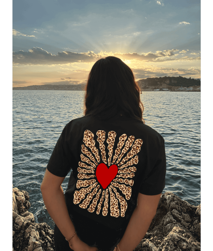 Femme portant un crop top noir 100% coton avec dans le dos personnalisation DTF coeur rouge au centre et traits léopard autour. Décor plage, coucher de soleil, mer, été