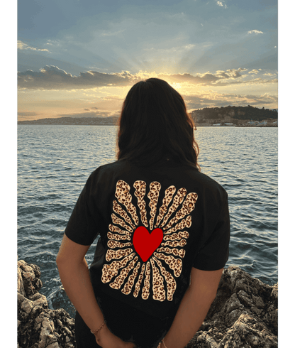 Femme portant un crop top noir 100% coton avec dans le dos personnalisation DTF coeur rouge au centre et traits léopard autour. Décor plage, coucher de soleil, mer, été
