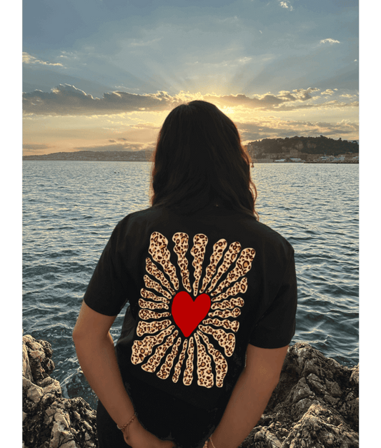 Femme portant un crop top noir 100% coton avec dans le dos personnalisation DTF coeur rouge au centre et traits léopard autour. Décor plage, coucher de soleil, mer, été