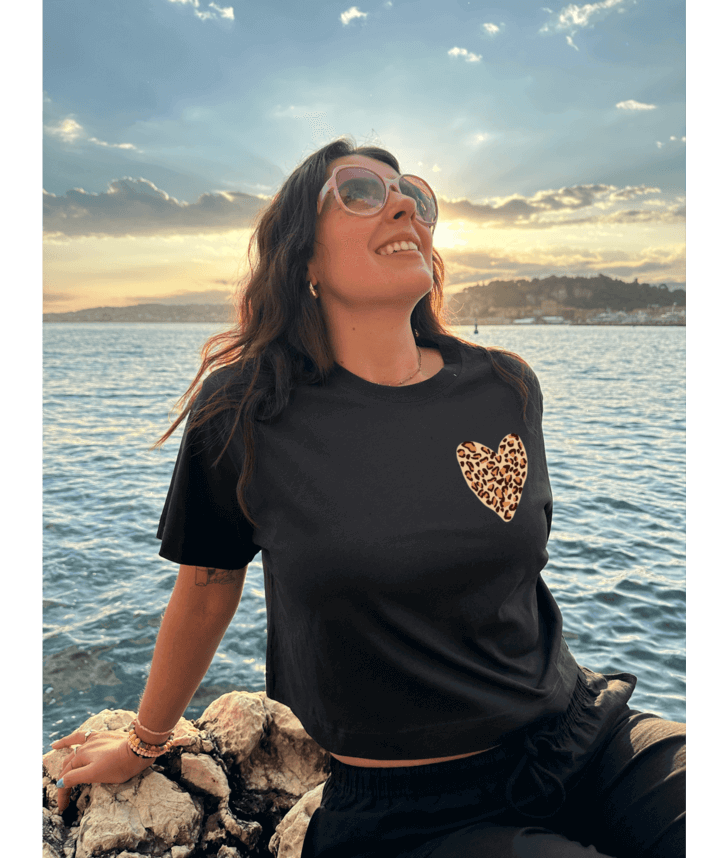 Femme portant un crop top noir 100% coton avec à l'avant un coeur léopard sur la poitrine et dans le dos personnalisation DTF coeur rouge au centre et traits léopard autour. Décor plage, coucher de soleil, mer, été