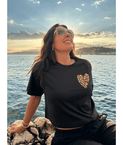 Femme portant un crop top noir 100% coton avec à l'avant un coeur léopard sur la poitrine et dans le dos personnalisation DTF coeur rouge au centre et traits léopard autour. Décor plage, coucher de soleil, mer, été