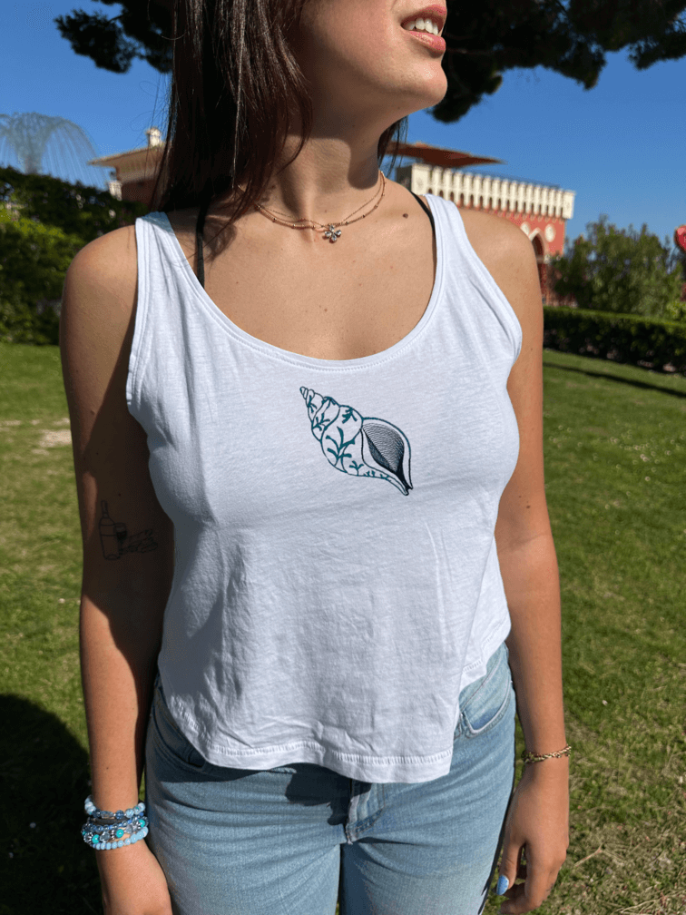 Débardeur blanc court, crop top, 100% coton. Personnalisation du débardeur grâce à des broderies. Il faut choisir parmi un choix de broderies dessinées à la main sur le thème de l’été sur la Côte d’Azur dans les Alpes Maritimes, 06. Il faut aussi choisir la position de la broderie entre à gauche côté cœur, à droite ou au milieu. Toutes les broderies sont faites à la commande selon la personnalisation choisie. Ici, il s’agit de la broderie coquillage bleu. Porté femme chateau Nice