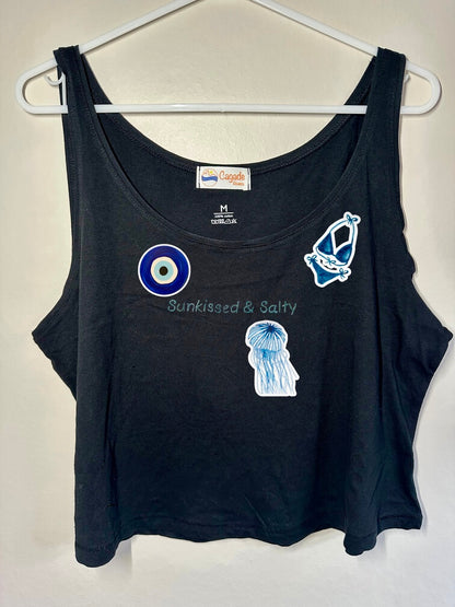 Débardeur noir court, crop top, 100% coton. Personnalisation du débardeur grâce à des broderies et du flocage DTF. Toutes les broderies sont faites à la commande. Ici, il s’agit de la broderie écriture "sunkissed & salty". et flocage DTF bleu de l'oeil de sainte lucie d'un maillot de bain femme bleu et d'une méduse bleue.