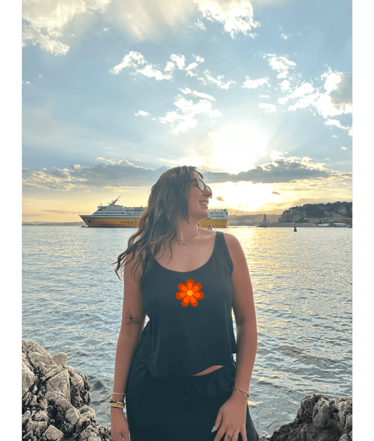 Débardeur noir court, crop top, 100% coton. Personnalisation du débardeur grâce à une technique de flocage, le DTF, pour une meilleure qualité d’impression et de tenue. La personnalisation est floquée au centre, en haut milieu. Ici, il s’agit de la personnalisation de DTF fleur orange seventies 70 milieu. Plage soleil été bord de l'eau mer coucher de soleil