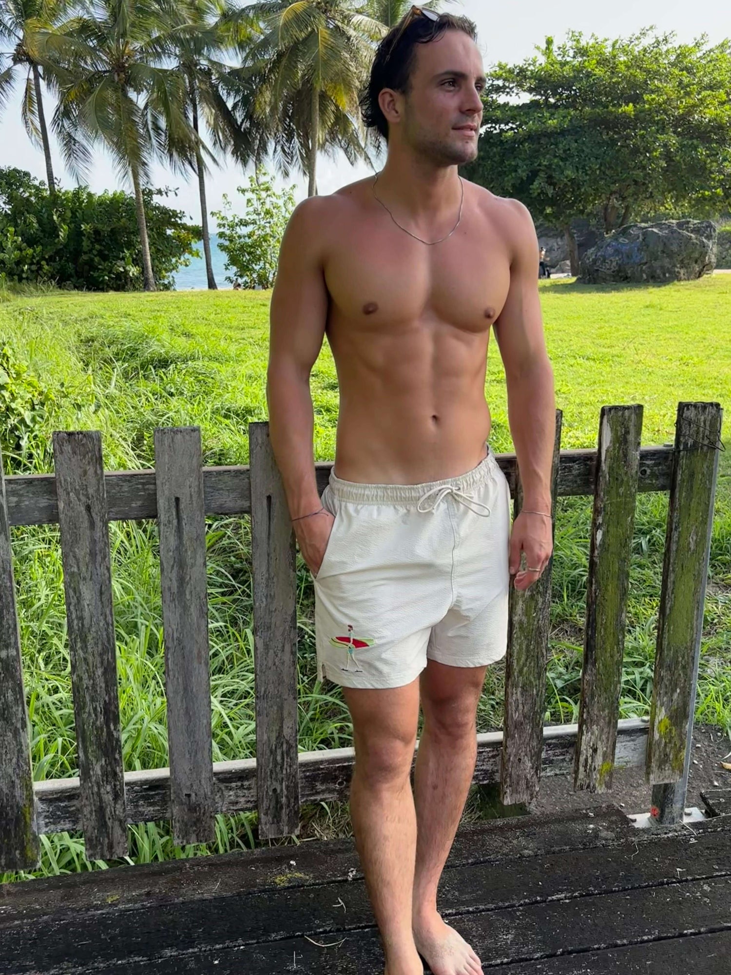 Mannequin homme portant maillot plage short beige avec broderie surf, surfeur avec sa planche de surf. Décor paradisiaque, Guadeloupe. 