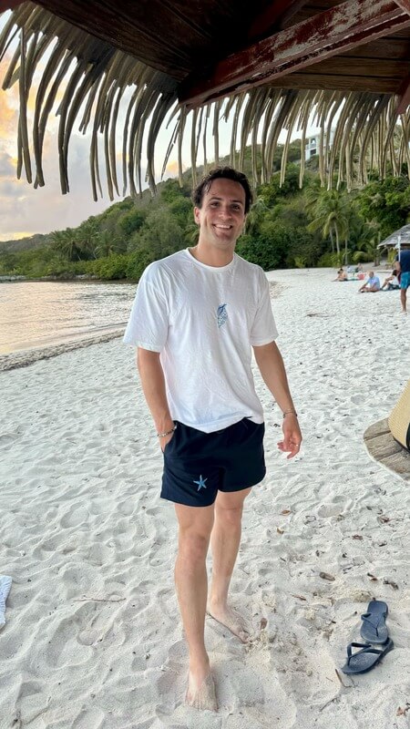 Mannequin, modèle homme portant un t-shirt oversize en conton personnalisé avec une broderie d'un coquillage bleu au centre et un maillot de bain plage bleu marine avec une personnalisation broderie étoile des mers bleu. Décor plage ile paradisiaque été 