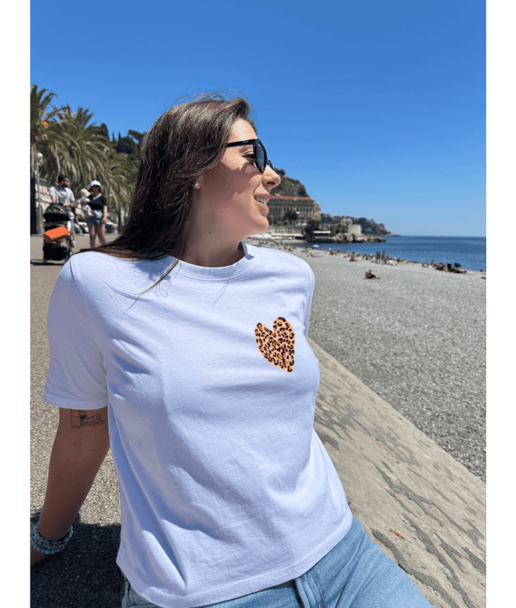 T-shirt oversize blanc, manche courte, 100% coton. Personnalisation du t-shirt grâce à une technique de flocage, le DTF, pour une meilleure qualité d’impression et de tenue. La personnalisation est floquée à l’avant au milieu en haut et à l’arrière en grand dans le dos. Ici, il s’agit de la personnalisation de DTF coeur léopard. Plage promenade des Anglais, Nice 06 été soleil.