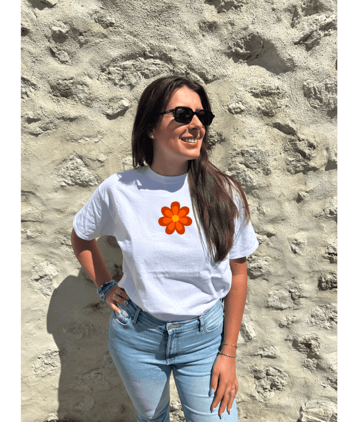 T-shirt oversize blanc, manche courte, 100% coton. Personnalisation du t-shirt grâce à une technique de flocage, le DTF, pour une meilleure qualité d’impression et de tenue. La personnalisation est floquée à l’avant au milieu en haut et à l’arrière en grand dans le dos. Ici, il s’agit de la personnalisation de DTF fleur orange style seventies. Porté par un modèle au bord de l'eau coucher de soleil mer plage été.