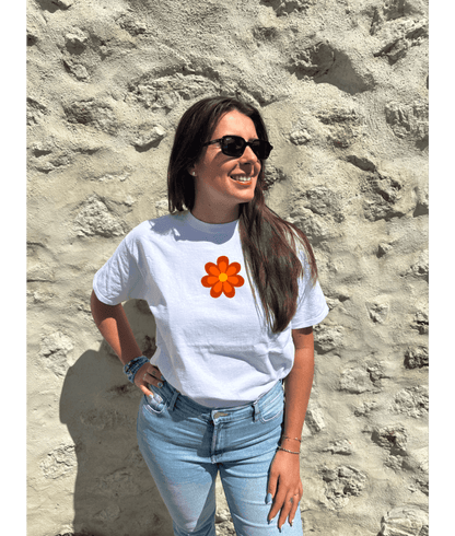 T-shirt oversize blanc, manche courte, 100% coton. Personnalisation du t-shirt grâce à une technique de flocage, le DTF, pour une meilleure qualité d’impression et de tenue. La personnalisation est floquée à l’avant au milieu en haut et à l’arrière en grand dans le dos. Ici, il s’agit de la personnalisation de DTF fleur orange style seventies. Porté par un modèle au bord de l'eau coucher de soleil mer plage été.