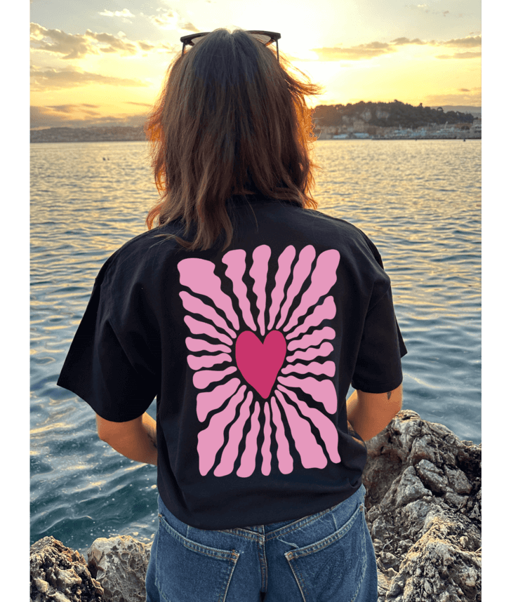 T-shirt oversize noir, manche courte, 100% coton. Personnalisation du t-shirt grâce à une technique de flocage, le DTF, pour une meilleure qualité d’impression et de tenue. La personnalisation est floquée à l’avant au milieu en haut et à l’arrière en grand dans le dos. Ici, il s’agit de la personnalisation de DTF coeur rose avant et arrière.
