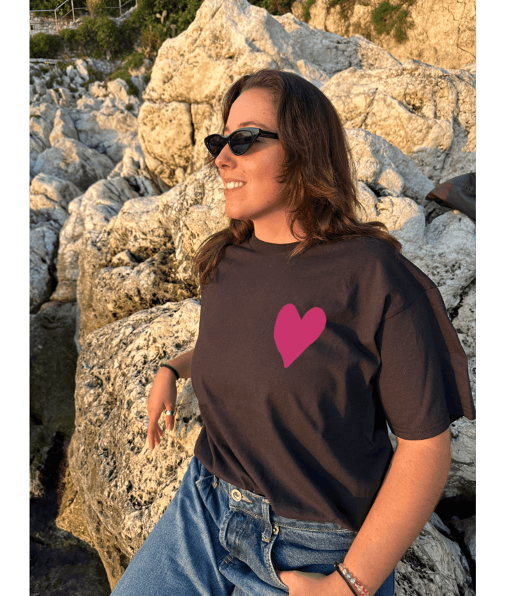 T-shirt oversize noir, manche courte, 100% coton. Personnalisation du t-shirt grâce à une technique de flocage, le DTF, pour une meilleure qualité d’impression et de tenue. La personnalisation est floquée à l’avant au milieu en haut et à l’arrière en grand dans le dos. Ici, il s’agit de la personnalisation de DTF coeur rose avant et arrière. Porté plage coucher de soleil mer