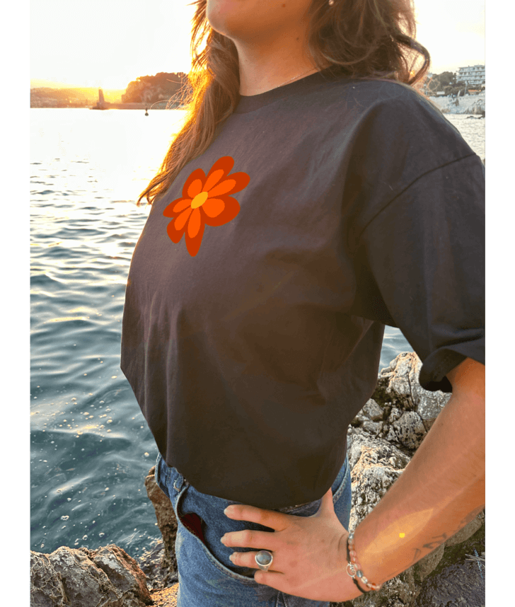 T-shirt oversize noir, manche courte, 100% coton. Personnalisation du t-shirt grâce à une technique de flocage, le DTF, pour une meilleure qualité d’impression et de tenue. La personnalisation est floquée à l’avant au milieu en haut et à l’arrière en grand dans le dos. Ici, il s’agit de la personnalisation de DTF fleur orange style seventies. Porté par un modèle au bord de l'eau coucher de soleil mer plage été.