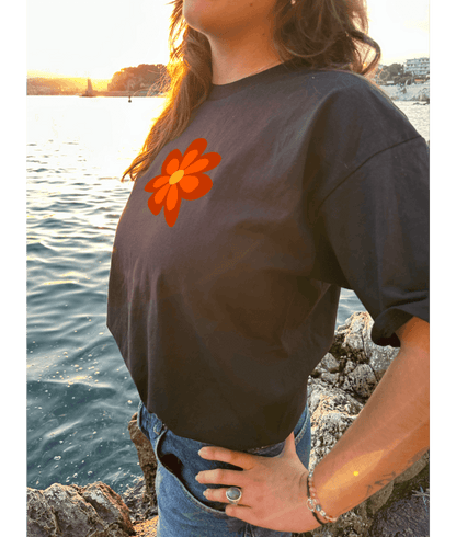 T-shirt oversize noir, manche courte, 100% coton. Personnalisation du t-shirt grâce à une technique de flocage, le DTF, pour une meilleure qualité d’impression et de tenue. La personnalisation est floquée à l’avant au milieu en haut et à l’arrière en grand dans le dos. Ici, il s’agit de la personnalisation de DTF fleur orange style seventies. Porté par un modèle au bord de l'eau coucher de soleil mer plage été.