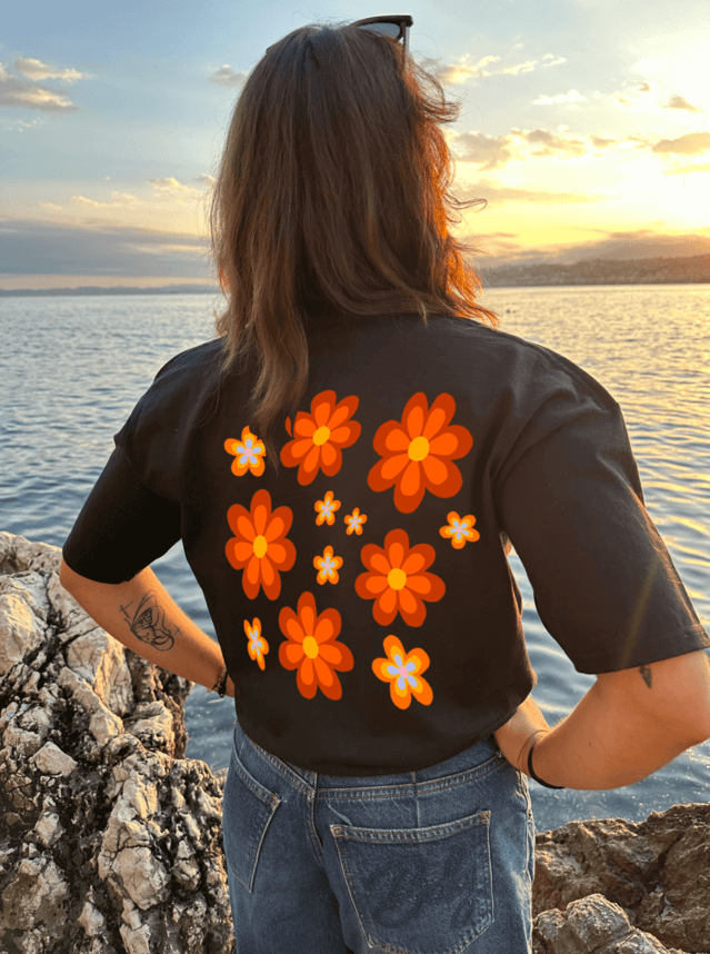 T-shirt oversize noir, manche courte, 100% coton. Personnalisation du t-shirt grâce à une technique de flocage, le DTF, pour une meilleure qualité d’impression et de tenue. La personnalisation est floquée à l’avant au milieu en haut et à l’arrière en grand dans le dos. Ici, il s’agit de la personnalisation de DTF fleur orange style seventies. Porté par un modèle au bord de l'eau coucher de soleil mer plage été.