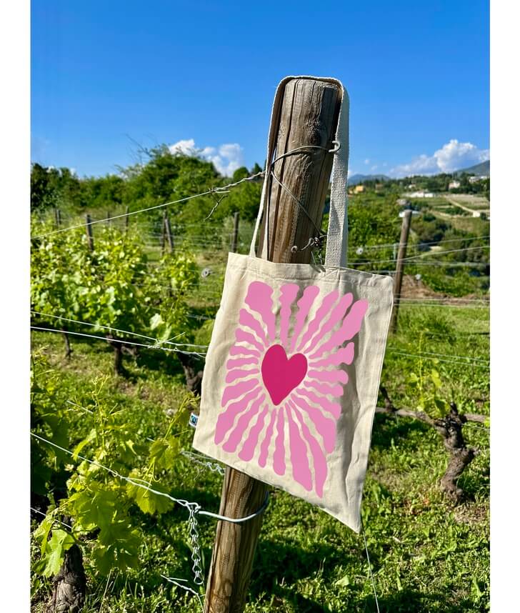 Tote bag, sac de course en coton. Personnalisation du débardeur grâce à une technique de flocage, le DTF, pour une meilleure qualité d’impression et de tenue. La personnalisation est floquée au centre, en haut milieu. Tout est fait à la commande selon la personnalisation choisie. Ici, il s’agit de la personnalisation de DTF, flocage coeur rose. Sac accroché dans les vignes, ciel bleu, verdure. 