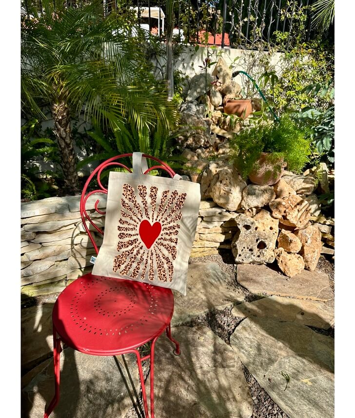 Tote bag, sac de course en coton. Personnalisation du débardeur grâce à une technique de flocage, le DTF, pour une meilleure qualité d’impression et de tenue. La personnalisation est floquée au centre, en haut milieu. Tout est fait à la commande selon la personnalisation choisie. Ici, il s’agit de la personnalisation de DTF, flocage coeur rouge et léopard. Sac posé sur une chaise rouge, au milieu des plantes. 