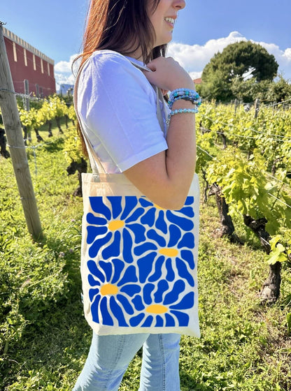 Tote bag, sac de course en coton. Personnalisation du débardeur grâce à une technique de flocage, le DTF, pour une meilleure qualité d’impression et de tenue. La personnalisation est floquée au centre, en haut milieu. Tout est fait à la commande selon la personnalisation choisie. Ici, il s’agit de la personnalisation de DTF, flocage fleur bleu. Décor dans les vignes, femme avec le sac sur l'épaule, ciel bleu, château. 