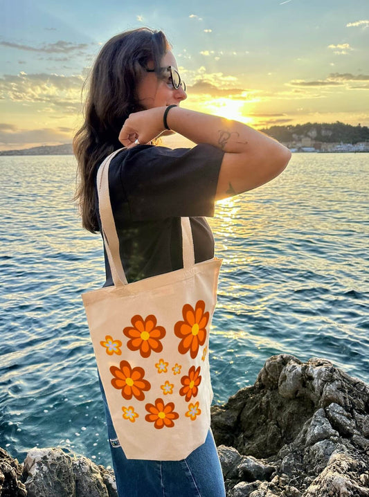 Tote bag, sac de course en coton. Personnalisation du débardeur grâce à une technique de flocage, le DTF, pour une meilleure qualité d’impression et de tenue. La personnalisation est floquée au centre, en haut milieu. Tout est fait à la commande selon la personnalisation choisie. Ici, il s’agit de la personnalisation de DTF, flocage fleur orange, inspiration seventies. Femme sac porté épaule, mer, coucher de soleil.
