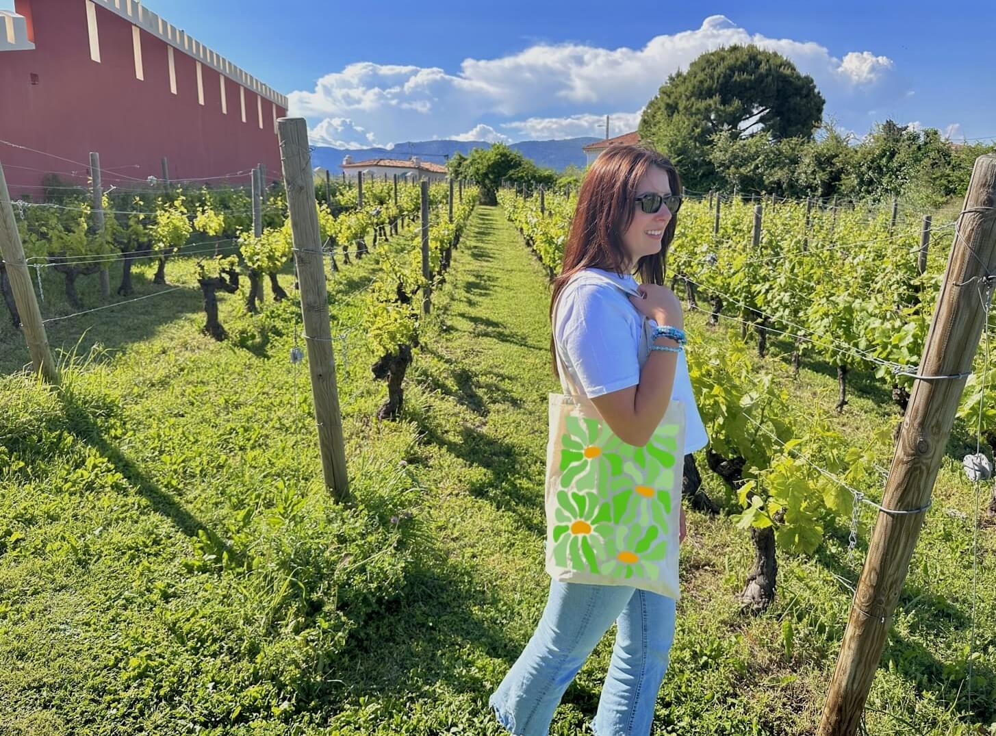 Tote bag, sac de course en coton. Personnalisation du débardeur grâce à une technique de flocage, le DTF, pour une meilleure qualité d’impression et de tenue. La personnalisation est floquée au centre, en haut milieu. Tout est fait à la commande selon la personnalisation choisie. Ici, il s’agit de la personnalisation de DTF, flocage fleur vert. Décor dans les vignes, femme avec le sac sur l'épaule, ciel bleu, château. 